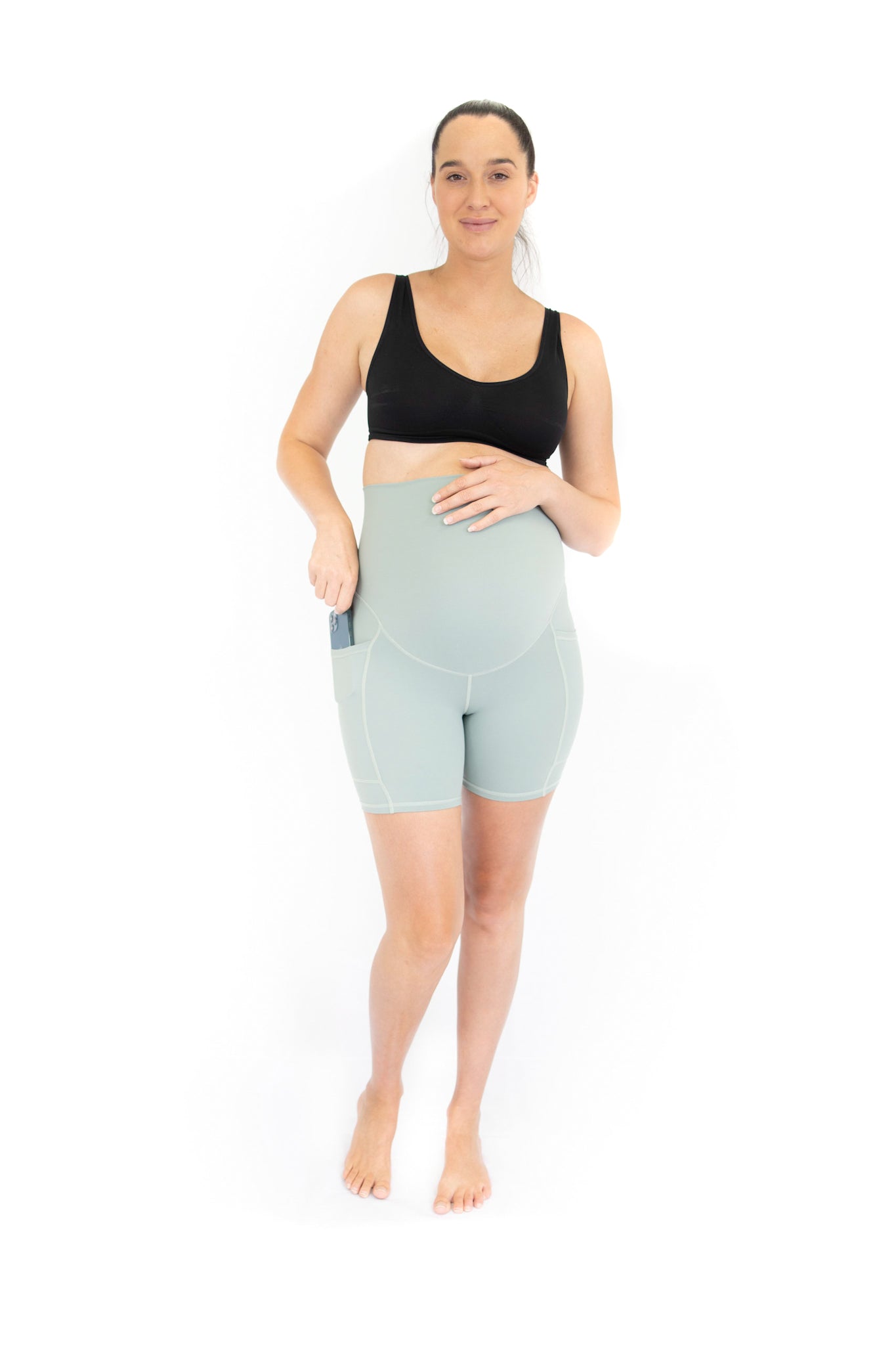 Woman in black sports bra and high-waisted mint green EmamaCo maternity bike shorts with pockets